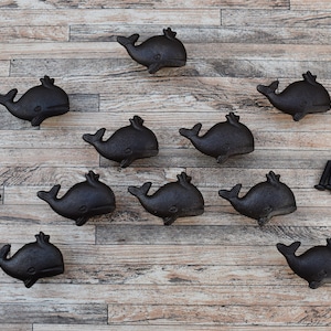May include: Nine black cast iron whale drawer pulls with screws. The whale pulls are arranged in a symmetrical pattern on a wood surface.