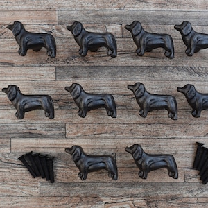 May include: Ten black cast iron dachshund dog drawer pulls with a rustic finish. The pulls are arranged in three rows on a wood surface. Each pull has a screw hole on the bottom.