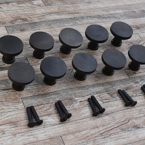 Könnte beinhalten: Ein Satz mit 15 schwarzen Metall-Schrankknöpfen mit runden Köpfen und einem kurzen Stiel. Die Knöpfe sind in drei Reihen zu fünf angeordnet. Das Set enthält außerdem 15 Schrauben zur Montage.