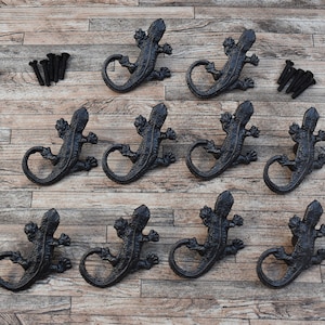Puede incluir: Diez ganchos de pared de lagarto de metal negro con tornillos. Los ganchos tienen forma de lagarto con la cola enroscada para formar un gancho. Los ganchos están dispuestos en tres filas sobre una superficie de madera.