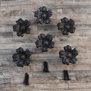 Op de afbeelding: Zes zwarte metalen bloemachtige ladegrepen met een decoratief ontwerp. De grepen zijn in een cirkel op een houten oppervlak gerangschikt met schroeven in het midden.