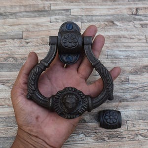 May include: Dark bronze door knocker with a detailed face design and floral accents. The knocker is held in a hand, showing its size and intricate details. Includes a matching strike plate. A decorative piece for any door.