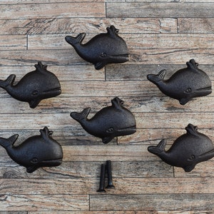 Puede incluir: Seis tiradores de cajón de ballena de hierro fundido negro con un acabado ligeramente texturizado. Los tiradores están dispuestos en círculo sobre un fondo de madera.