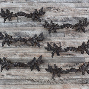 Peut inclure: Six poignées de tiroir en fonte noire avec un motif de feuilles et de volutes. Les poignées sont disposées en deux rangées de trois sur un fond en bois.