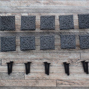 May include: A set of ten square, dark gray cabinet knobs with an ornate, swirling pattern. The knobs are arranged in two rows, with a set of black screws below them. The knobs are made of metal and are likely for furniture.