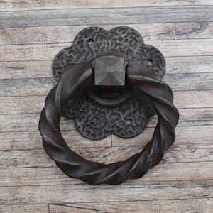 May include: A dark brown, cast iron door knocker with a twisted handle. The knocker has a decorative floral backplate and a square center piece. The door knocker is designed to be mounted on a door.