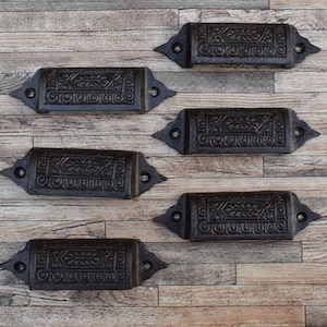 May include: Five antique black cast iron drawer pulls with ornate designs. The pulls are arranged in a row on a wood surface.