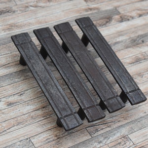 Può includere: Quattro maniglie per mobili marrone scuro con un design rettangolare. Ogni maniglia ha una superficie strutturata ed è montata su uno sfondo di legno. Le maniglie sono distanziate uniformemente e sembrano essere in metallo.