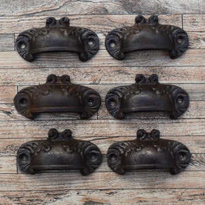 May include: Six antique-style black cast iron drawer pulls with ornate detailing. The pulls are arranged in two rows of three on a light brown wood background.
