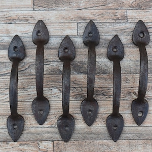 Könnte beinhalten: Sechs rustikale schwarze Metall-Türgriffe mit spitzen Enden. Die Griffe sind in einer Reihe auf einer Holzoberfläche angeordnet.