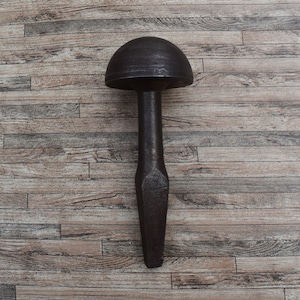 Puede incluir: Una herramienta de metal oscuro de aspecto antiguo, con una cabeza redondeada y un mango largo y cónico. La herramienta probablemente se utiliza para dar forma o formar materiales. El fondo es una superficie de madera.
