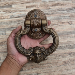May include: An ornate brass door knocker with a detailed design featuring a face and floral motifs. The knocker is mounted on a wooden door.