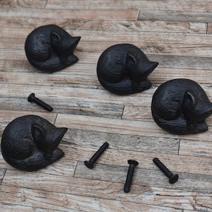 Könnte beinhalten: Vier schwarze Guss-Katzen-Türgriffe mit einem schlafenden Katzen-Design. Die Griffe befinden sich auf einer Holzoberfläche mit vier schwarzen Schrauben.