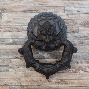 Op de afbeelding: Een sierlijke zwarte deurklopper van gietijzer met een gedetailleerd ontwerp met een centraal gezicht en een ringvormige handgreep. De klopper is gemonteerd op een achtergrond van houten planken.