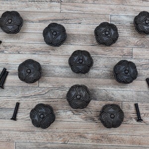 May include: Ten black metal flower-shaped cabinet knobs with a decorative design. The knobs are arranged in a grid pattern on a wooden surface. Each knob comes with two screws.