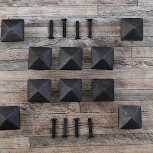May include: A set of eight black metal pyramid-shaped decorative nail heads or furniture studs arranged in rows, accompanied by eight matching black metal screws. Each stud features a square base and a pointed top, with a dark, textured finish.