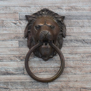 Peut inclure: Un heurtoir de porte en bronze en forme de tête de lion avec une bague circulaire. Le heurtoir est monté sur une surface en bois.