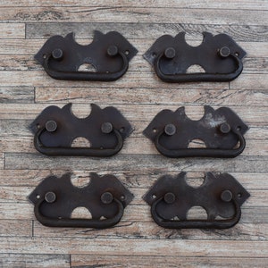 May include: Six antique-style metal drawer pulls with a decorative design. The pulls are dark brown and have a rustic finish.