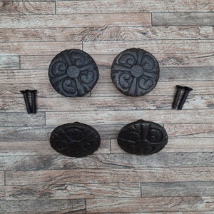 Può includere: Quattro pomelli per cassetti rotondi in ghisa nera con un design floreale decorativo. I pomelli sono rotondi e hanno un centro leggermente rialzato. Sono mostrati su una superficie in legno con due viti.