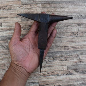 May include: A black metal anvil with a pointed tip, held in a person's hand. The anvil is a traditional blacksmithing tool used for shaping metal.