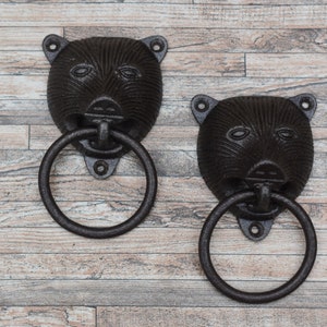 Puede incluir: Dos llamadores de puerta de hierro fundido negro en forma de cabeza de oso con anillos redondos. Los llamadores están montados sobre un fondo de madera.