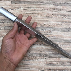 May include: A silver-colored metal mandrel with a cylindrical handle section that transitions into a long, tapered, multi-sided shaft. The tool features a smooth, reflective surface and a pointed end, designed for shaping or sizing circular items.