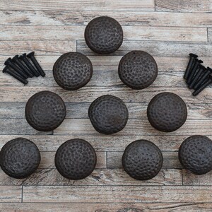 May include: Ten black metal cabinet knobs with a hammered finish. The knobs are arranged in a triangle shape on a wooden surface.