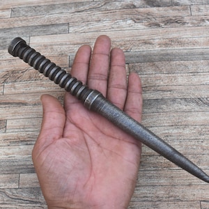 May include: A metal wand with a textured, ridged handle and a tapered, pointed end. The wand is held in a hand, showcasing its design and craftsmanship. The wand's color is a muted gray, suggesting a vintage or antique aesthetic.