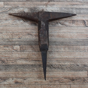 Puede incluir: Un yunque vintage, una herramienta de herrero, está centrado sobre un fondo de tablones de madera. El yunque es de color gris oscuro con un cuerpo cónico y una parte superior plana y rectangular con dos cuernos. La base es una espiga larga y puntiaguda.