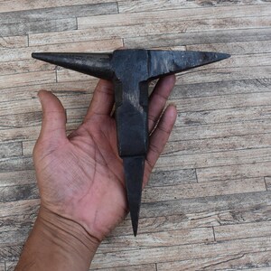 Pode incluir: Uma bigorna de metal preto com uma ponta pontiaguda, segurada na mão contra um fundo de grão de madeira.