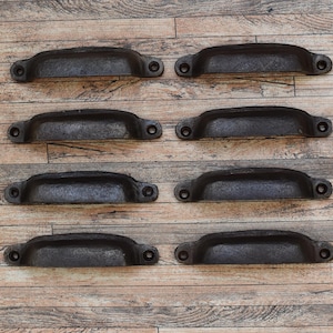 May include: Eight antique-style, black cast iron drawer pulls on a wooden surface. Each pull has a curved, cup-like design with two mounting holes. The pulls are evenly spaced on the wooden surface.