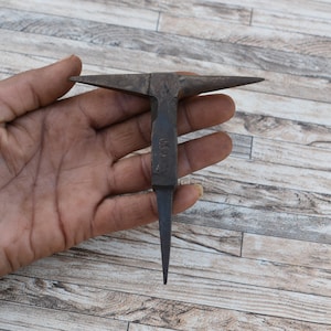 May include: A small, antique metal anvil with a pointed tip and a flat top. The anvil is held in a hand, and the background is a wooden surface.