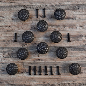 Könnte beinhalten: Zehn schwarze Metall-Schrankknöpfe mit gehämmerter Oberfläche und runder Form. Die Knöpfe sind symmetrisch auf einer Holzoberfläche angeordnet. Das Bild zeigt auch 12 schwarze Schrauben.
