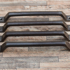 Puede incluir: Cuatro tiradores de cajón de metal negro con forma rectangular y bordes redondeados. Los tiradores están dispuestos en fila sobre una superficie de madera.