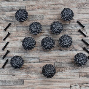 May include: Ten black cast iron drawer pulls with a floral design. The pulls are arranged in a pattern on a wood surface.