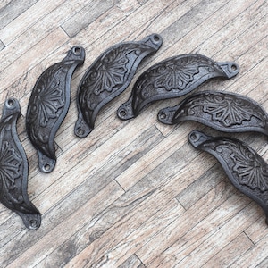 May include: A collection of seven dark gray, cast iron cabinet pulls. Each pull has a curved, cup-like shape with an ornate floral design. The pulls are arranged on a light brown, wood-grain surface.
