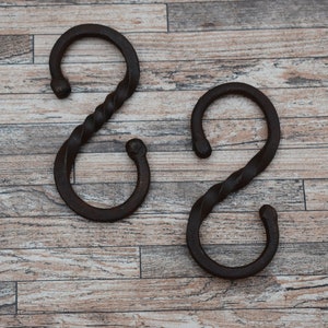 May include: Two black metal S-shaped hooks with a twisted design. The hooks are on a woodgrain surface.