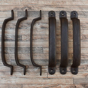 Puede incluir: Seis tiradores de cajón de metal negro con un diseño curvo. Los tiradores tienen un acabado rústico y están dispuestos en una fila sobre una superficie de madera.