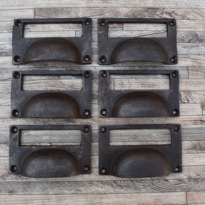 May include: Six antique-style drawer pulls with a rectangular label plate and a rounded, curved handle. The pulls are made of dark metal with a distressed finish.