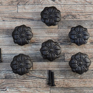 Può includere: Sei tiretti per cassetti in ghisa nera con un design floreale. I tiretti sono disposti in cerchio su una superficie in legno.