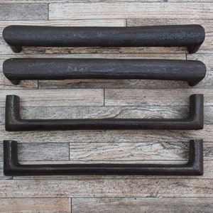 Può includere: Quattro maniglie per cassetti in metallo nero con forma rettangolare arrotondata. Le maniglie sono disposte verticalmente su una superficie in legno.