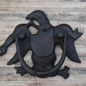 Pode incluir: Um batedor de porta de ferro fundido preto em forma de águia com as asas abertas. O batedor tem um anel para bater.