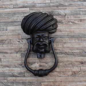May include: A black cast iron door knocker shaped like a person's head wearing a turban. The knocker has a detailed face with a mustache and beard.
