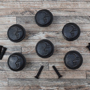Könnte beinhalten: Sechs schwarze Schrankknöpfe aus Gusseisen mit einem Halbmond-Design. Die Knöpfe sind in einem Kreis auf einer Holzoberfläche angeordnet. Zwei Sätze Schrauben sind ebenfalls enthalten.