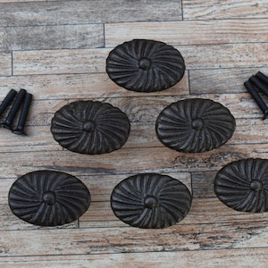 Op de afbeelding: Zes zwarte ovale kastknoppen met een gestructureerd, stralend ontwerp. De knoppen worden vergezeld door zwarte schroeven, allemaal gerangschikt op een lichtbruin houten oppervlak. Deze decoratieve knoppen zijn geschikt voor meubels.