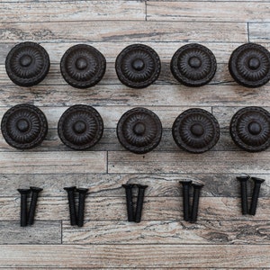 May include: Ten black metal cabinet knobs with a decorative design and eight black screws. The knobs and screws are arranged on a wooden surface.