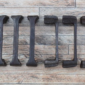 Könnte beinhalten: Ein Set aus sechs dunkelgrauen Metallgriffen mit einer texturierten, verwitterten Oberfläche. Drei Griffe sind vertikal und drei horizontal angeordnet, was ein schlichtes, rechteckiges und funktionales Design für Möbelstücke darstellt.