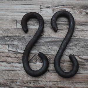 Może przedstawiać: Dwa rdzawe metalowe haczyki w kształcie litery S, prawdopodobnie używane do podnoszenia lub mocowania przedmiotów, umieszczone są obok siebie na drewnianej powierzchni.
