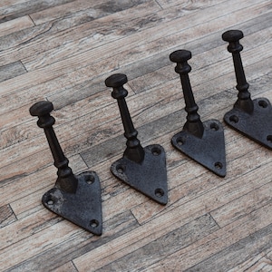 May include: Four black metal hooks with a triangular base and a decorative design. The hooks are arranged in a row on a wooden surface.