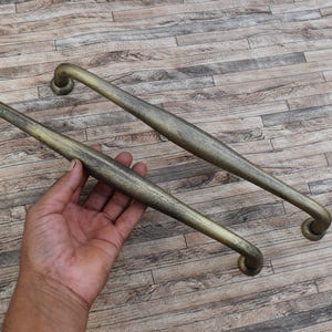 Könnte beinhalten: Zwei antike Messing-Schrankgriffe mit geschwungenem Design. Die Griffe haben eine warme, gealterte Messingfarbe und eine leicht strukturierte Oberfläche. Die Hardware ist für eine einfache Installation konzipiert und verleiht einen Vintage-Touch.